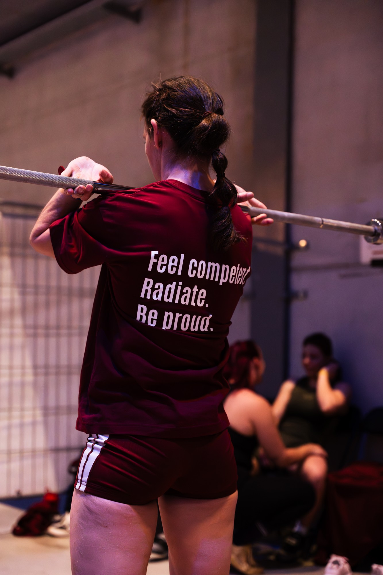 Een atleet met de stang op de schouders, met op haar shirt Feel competent. Radiate. Be proud.